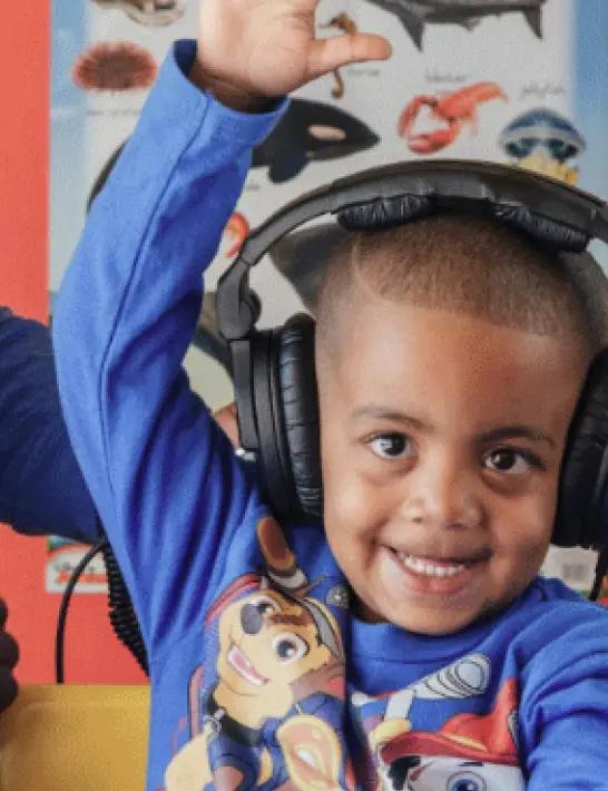 Smiling little boy in a classroom, taking a hearing test and raising his hand.